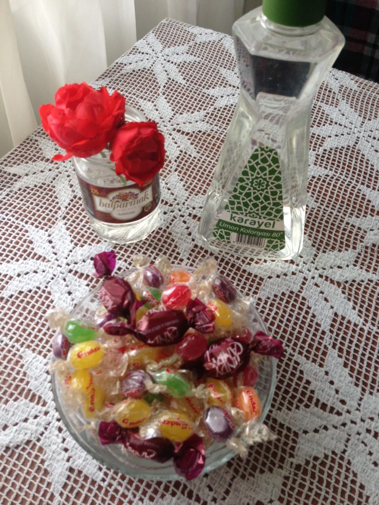 A bowl of multi-colored candies sits in front of two red flowers in a jar of water and a bottle of limon kolonya. They are placed on a tabletop with a white lace covering and white curtains peek out behind the table. These are preparations for visitors during Seker Bayram that marks the end of Ramadan in Turkey.