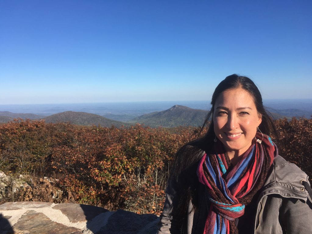 Malia Yoshioka stands on a lookout from Shenandoah Valley's Skyline Drive. In the background is a bright blue sky and an expanse of trees turning orange and gold with fall foliage.