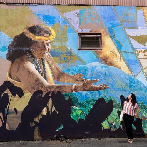 Malia Yoshioka standing in front of a giant graffiti wall of a Hawaiian woman reaching out
