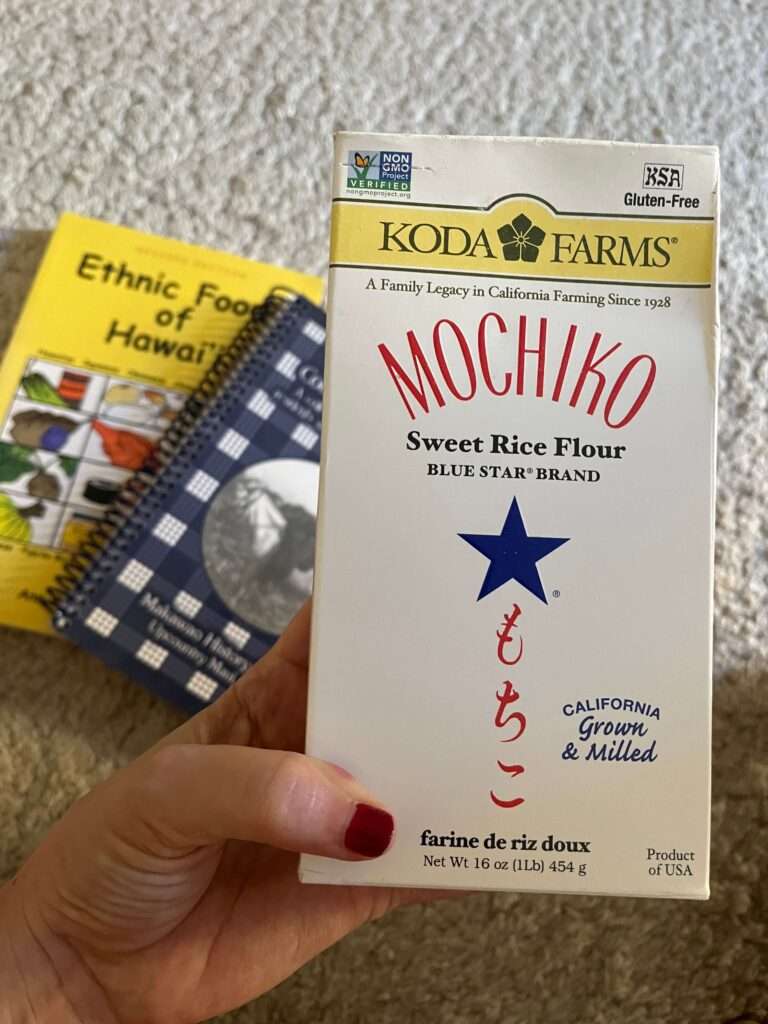 A hand holding a box of Koda Farms Blue Star Brand Mochiko (sweet rice flour). The box also says "A family Legacy in California Farming since 1928" and "California Grown & Milled". In the background there are also spiral bound cookbooks from Makawao in Maui, Hawaii and Ann Konda Corum's Ethnic Foods of Hawaii book.