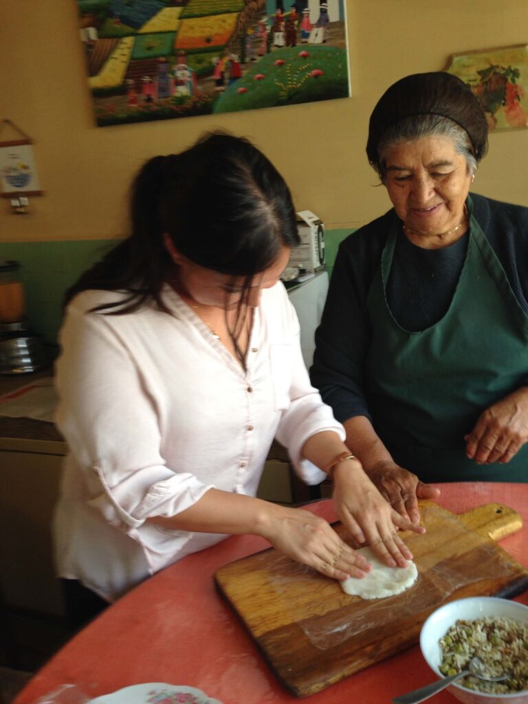 Two ladies in a kitchen in Quito, Ecuador look over a table during an Ecuadorian cooking class in Quito. Malia Yoshioka pressed down a circle of dough with the instructor watching over her shoulder. A bowl of filling sits in front of them on the table and colorful folk paintings adorn the walls behind.