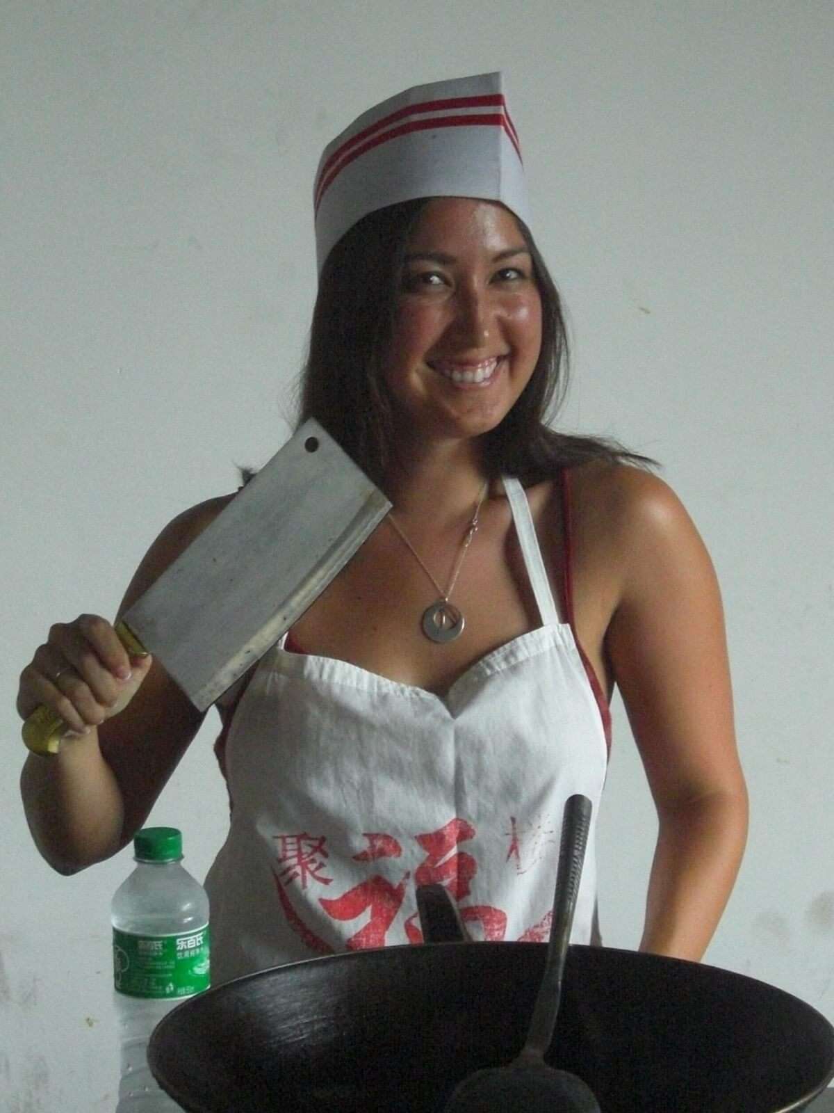 Malia Yoshioka holds a cleaver and stands dressed in an apron and white paper hat in a cooking class in Yangsuo, China. There is a wok and spatula in front of her.