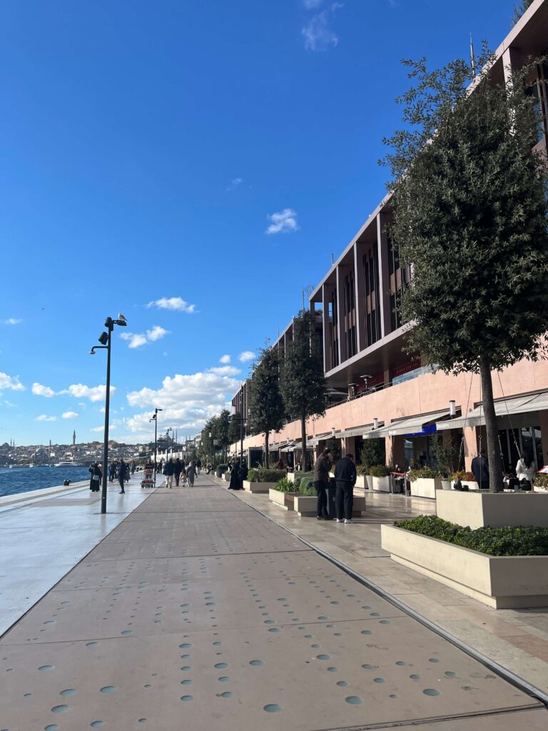 The walkway in front of the Galaport shopping complex in Istanbul is a 1.2km stretch along the Bosphorus strait on the left side of the photo. To the right are 2 levels of shops and restaurants, a few concrete planters with trees in front of them. It's a bright blue sky day and there are no cruise ships to block the sea view.