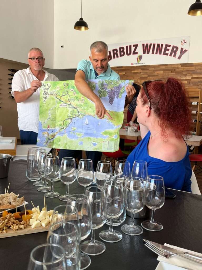 Akin Gurbuz, owner of Gurbuz Winery in Tekirdag, Turkiye leads a Turkish wine tasting. A man helps him to hold up a map of the region around Istanbul and Tekirdag to show the locations where the grapes were grown. Andrea Lemieux, the author of the Essential Guide to Turkish Wine, sits at the table facing Akin with her back to the camera. On the table, 8 wine glasses per person are set empty and ready for the wines to be poured, with cheese and nuts and dried fruits on cutting boards to accompany the tasting.