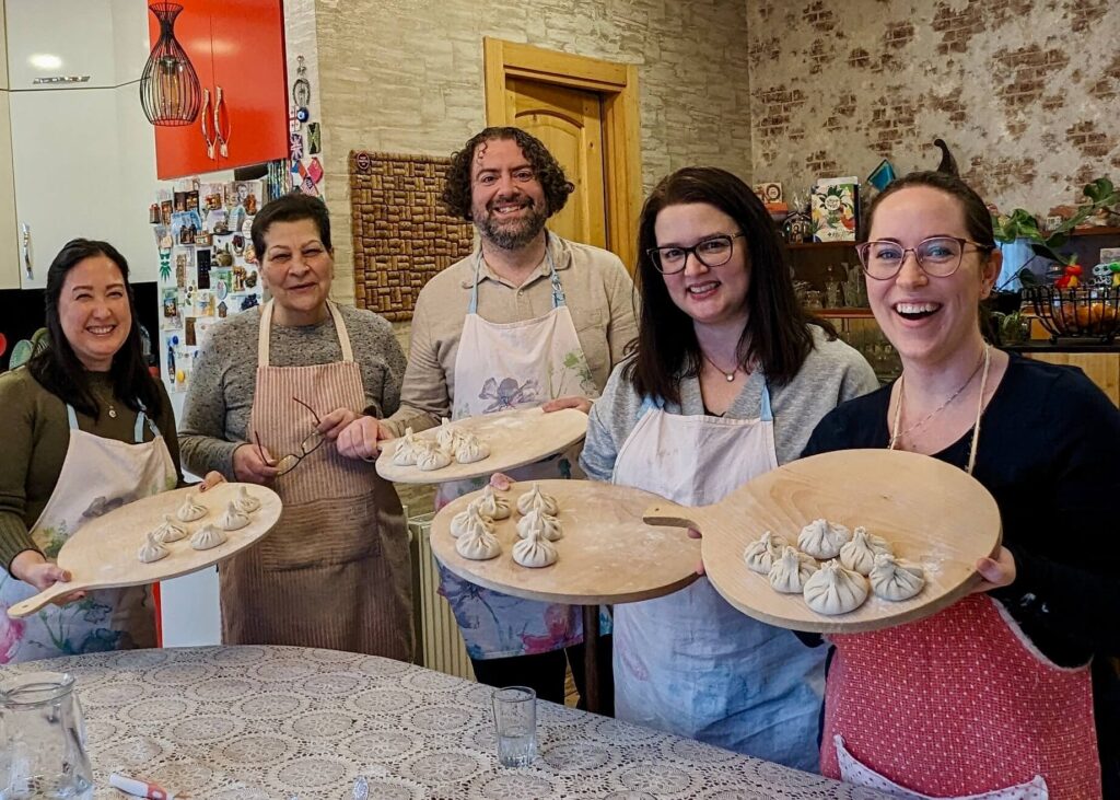 Four friends and their cooking instructor at a cozy living room and kitchen in Tbilisi, Georgia. The students are dressed in aprons and hold circular boards with handmade khinkali ready to be cooked. This is a perfect culinary experience for foodies in Georgia.