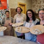 Four friends and their cooking instructor at a cozy living room and kitchen in Tbilisi, Georgia. The students are dressed in aprons and hold circular boards with handmade khinkali ready to be cooked.