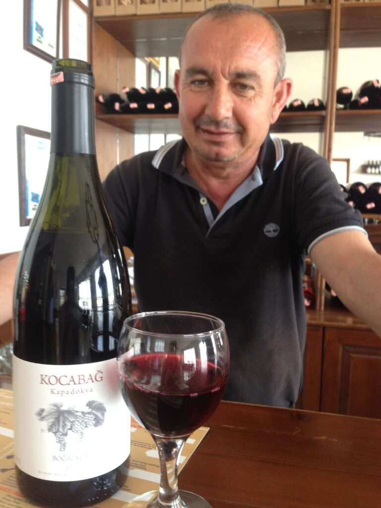 Mehmet Erdogan stands behind the tasting room bar at Kocabag winery in Urgup, Cappadocia, Turkiye. There is a glass of red wine and a red wine bottle that says Kocabag Kapadokya - its a turkish wine grape called Bogazkere which translates to "throat burner" because of the tannins.