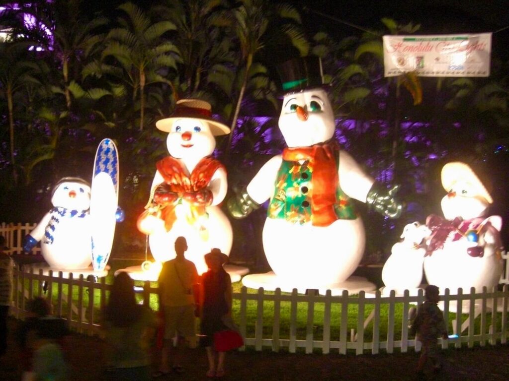 A snow family (including dad in an aloha shirt and top hat and the youngest snowman with a surfboard) is set up during Honolulu City Lights holiday display and surrounded by a fence with families visiting the holiday decorations.