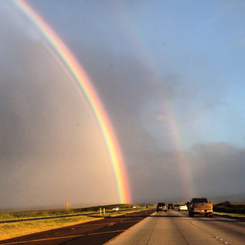 A double rainbow arches over Haleakala Highway in Maui, Hawaii. The main rainbow on the left glows so strongly because there's a cloud of rain behind it, and the right one is lighter. Behind both you can see a blue sky over the dormant volcano, Haleakala. four lanes of traffic span the highway below as residents commute home.