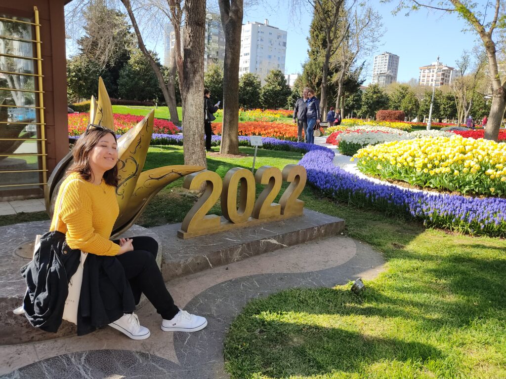 the author, Malia Yoshioka sits in Goztepe Park in Istanbul next to a sign saying 2022 during the springtime Tulip Festival. She's wearing a yellow sweater and brightly colored landscapes of tulips are visible across the park in the background, with apartment building in the distance. Goztepe Park on the Asian side of Istanbul is one of the popular places to see the Istanbul Tulip Festival each spring.