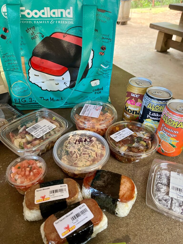 A spread of four types of poke, three spam musubi and juice and canned drinks - a local picnic feast - in front of a bag from Foodland supermarket with a drawing of a spam musubi. It was for a picnic at Iao Valley on Maui to compare different types of poke.