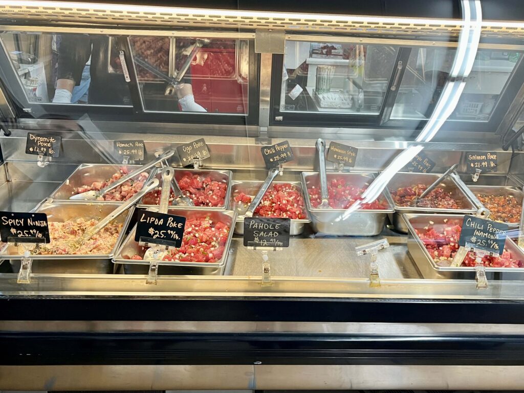 A view of the poke counter at Kaohu Store in Wailuku, Maui. Various types of poke are displayed in silver trays including spicy ninja, lipoma poke, pahole salad, chili pepper inamona, dynamite poke, ogo poke, and more. This is the author's favorite poke on Maui.