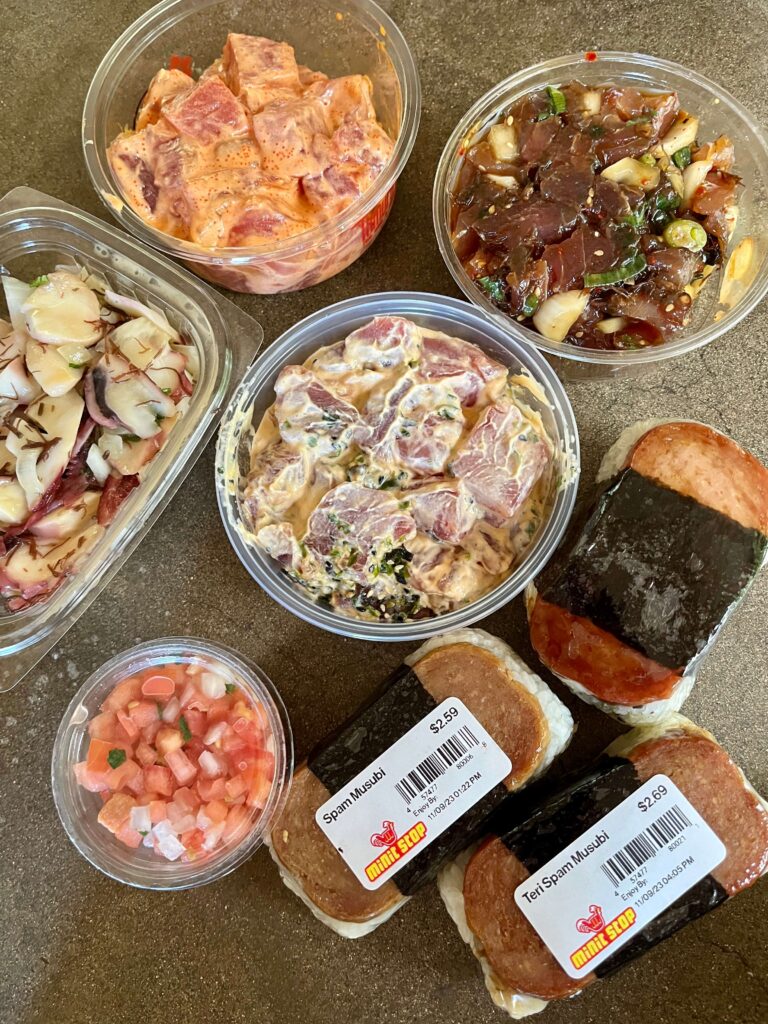 A top down look at four different kinds of poke in containers lined up to compare for Central Maui's best poke. Sesame Tako poke from Takamiya's, Shoyu ogo poke from Oki's Seafood, Spicy Ahi poke from Tamura's and Spicy Ninja poke from Kaohu Store, along with a small container of lomi lomi salmon and three spam musubis. Local Hawaiian food at its best!