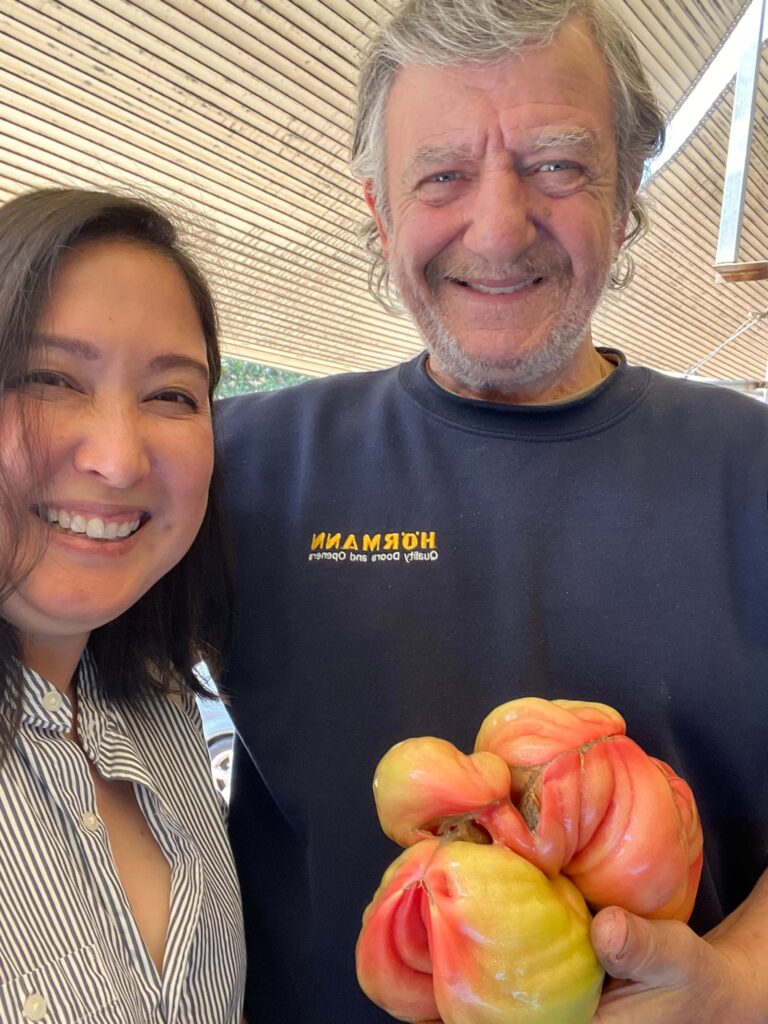 Malia Yoshioka smiles beside a local farmer at the Kalamata open market, holding a large, uniquely shaped heirloom tomato known in Greek as “fat and curly.” The vibrant tomato features swirls of red, pink, and yellow, showcasing the region’s rich agricultural produce.