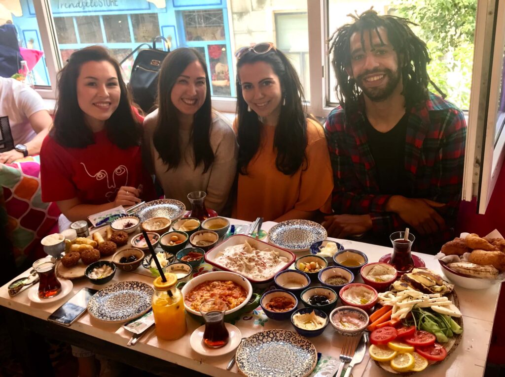 Author Malia Yoshioka and friends enjoying a Turkish-Lebanese breakfast feast at Arada Café (Arada Beyrut) in Istanbul, with a colorful spread of small dishes including labneh, hummus, muhammara, olives, eggs, cheeses, fresh vegetables, falafel, and Turkish tea, blending Middle Eastern and Turkish breakfast traditions in a vibrant and instagram-worthy café setting.