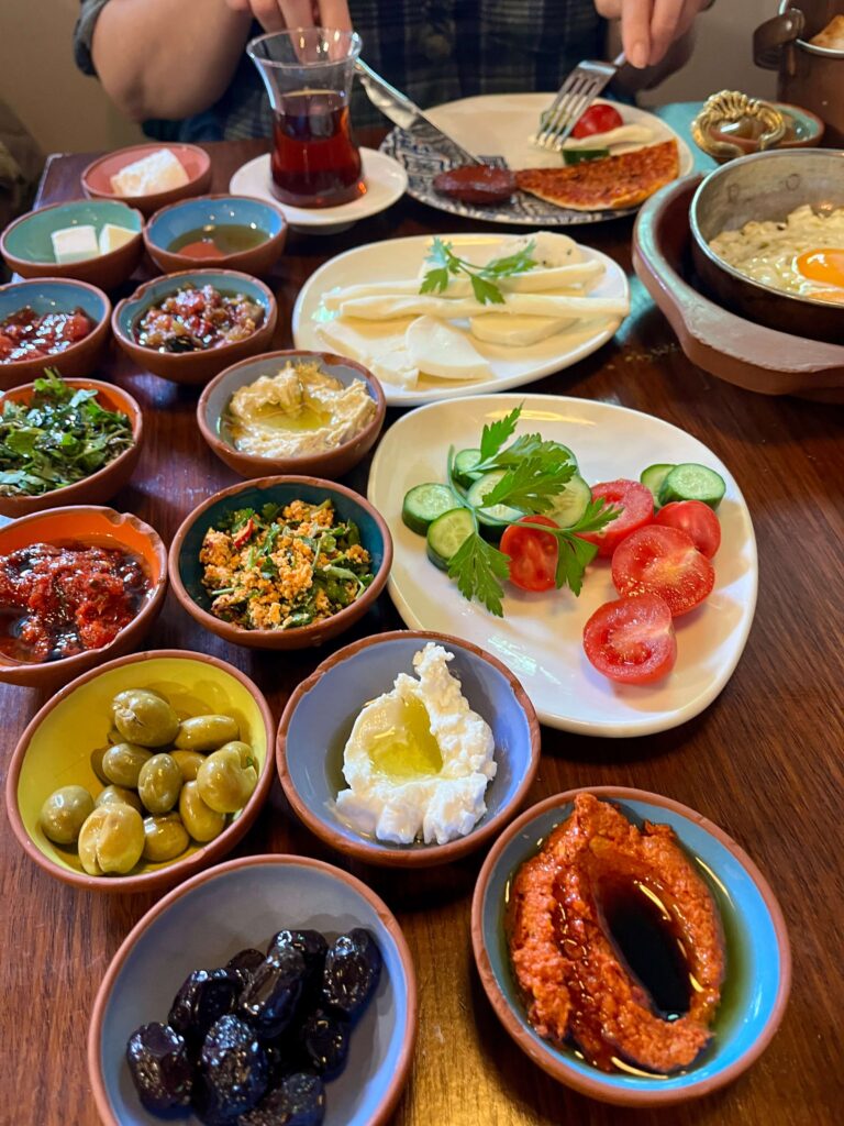 Traditional Turkish breakfast in Istanbul at Doğacıyız, showcasing specialties from Hatay/Antakya, including small green olives native to the region, creamy clotted cheese with olive oil, muhammara, assorted jams, herbs, and fresh veggies, served with sahanda yumurta (fried egg in a pan) and Turkish tea in curved glasses.