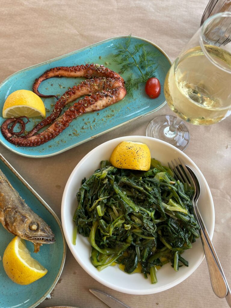A Greek seafood meal from The Blue Shrimp in Kalamata, Greece is served on a paper-covered table, featuring grilled octopus garnished with herbs and dill on a blue plate, a side of steamed greens drizzled with olive oil and lemon in a white bowl, and a whole grilled fish accompanied by lemon wedges. A glass of white wine completes the spread, suggesting a fresh, coastal dining experience just steps away from the Kalamata Open Market and its lovely fish counters.