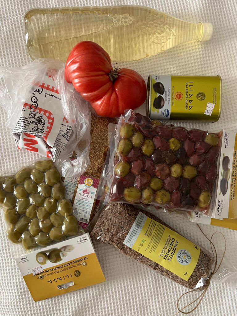 A selection of goods from the Kalamata open market displayed on a white surface, including a large heirloom tomato, bottled white wine, Kalamata olives, green olives stuffed with lemon, sesame pasteli, barley rusks, and a tin of Iliada brand olives. The haul highlights fresh, regional products typical of the Peloponnesian market scene.