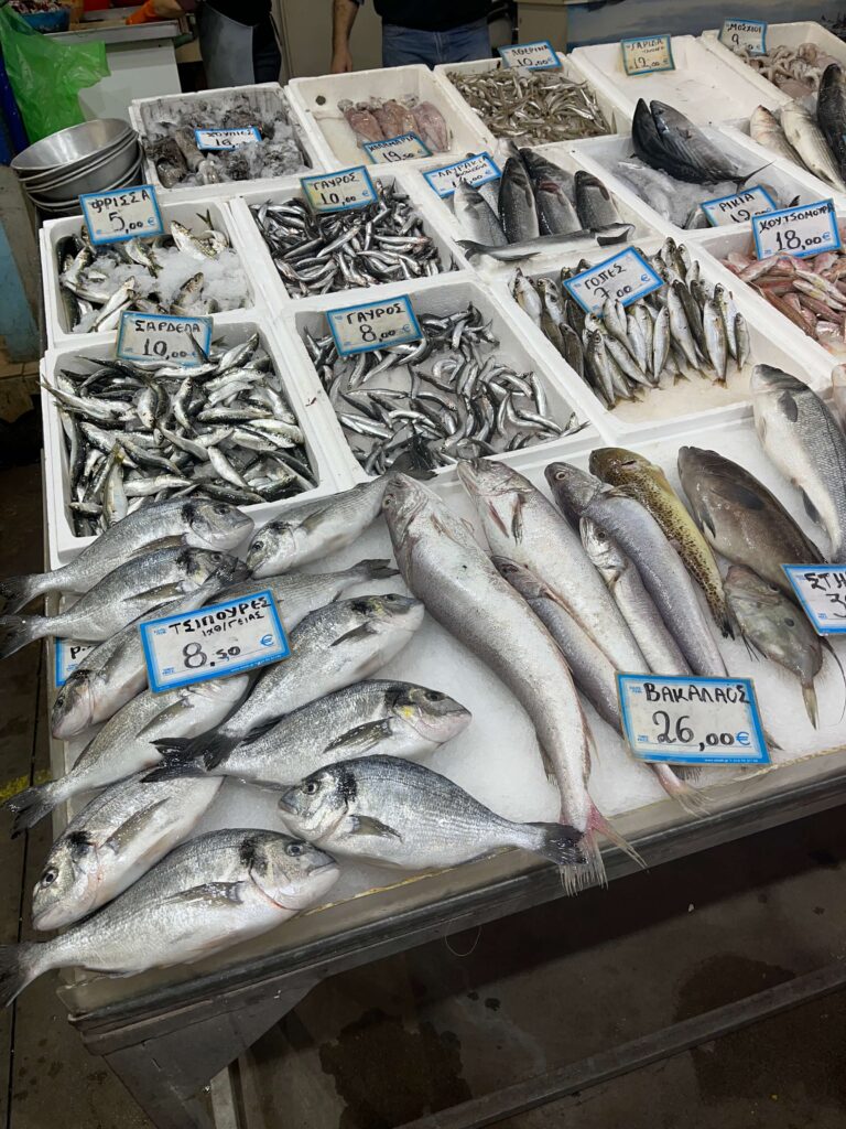 A wide selection of fresh seafood on ice at the Kalamata open market, including sardines, anchovies, gilt-head bream (τσιπούρες), cod (μπακαλάος), and shrimp, each labeled with handwritten Greek signs and prices in euros. The fish stall captures the variety and abundance of the region’s coastal offerings.