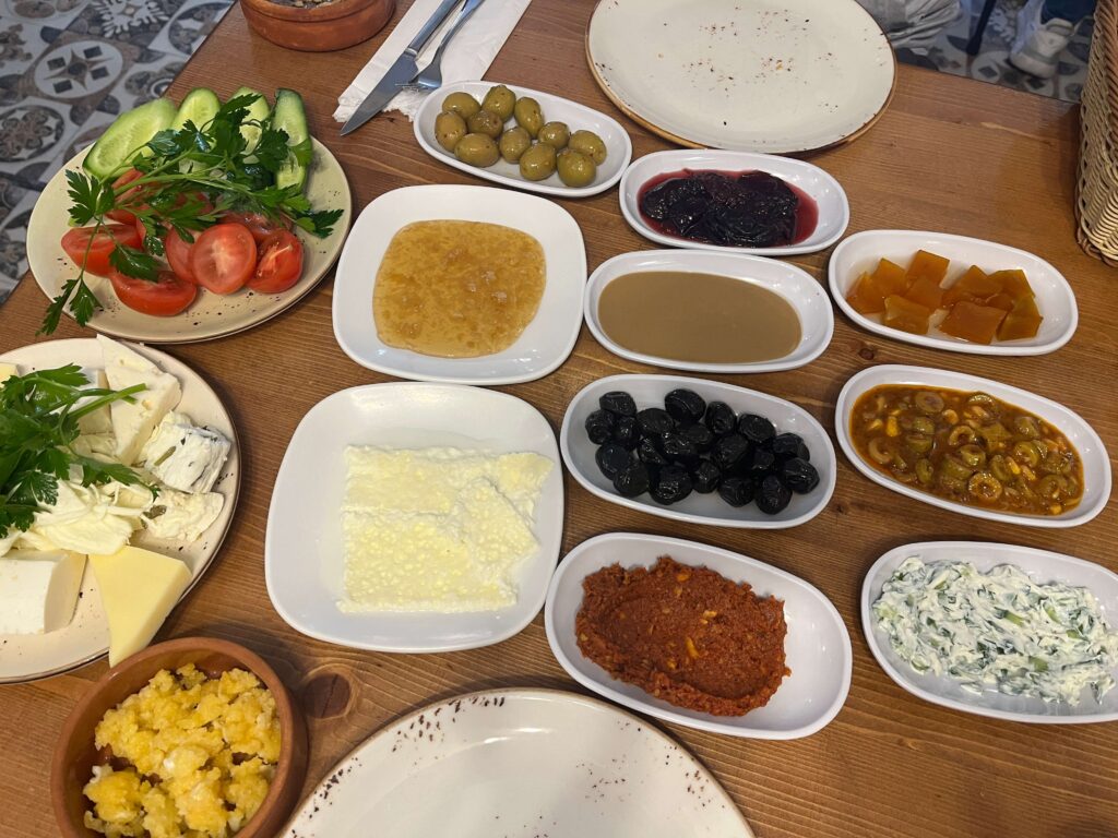 Traditional Turkish breakfast spread at Van Kahvaltı Evi in Istanbul, featuring a variety of cheeses, black and green olives, fresh tomatoes and cucumbers, honey, clotted cream, jams, tahini, herbed yogurt dip, and regional specialties like menemen and muhammara, all served in small white dishes on a rustic wooden table.