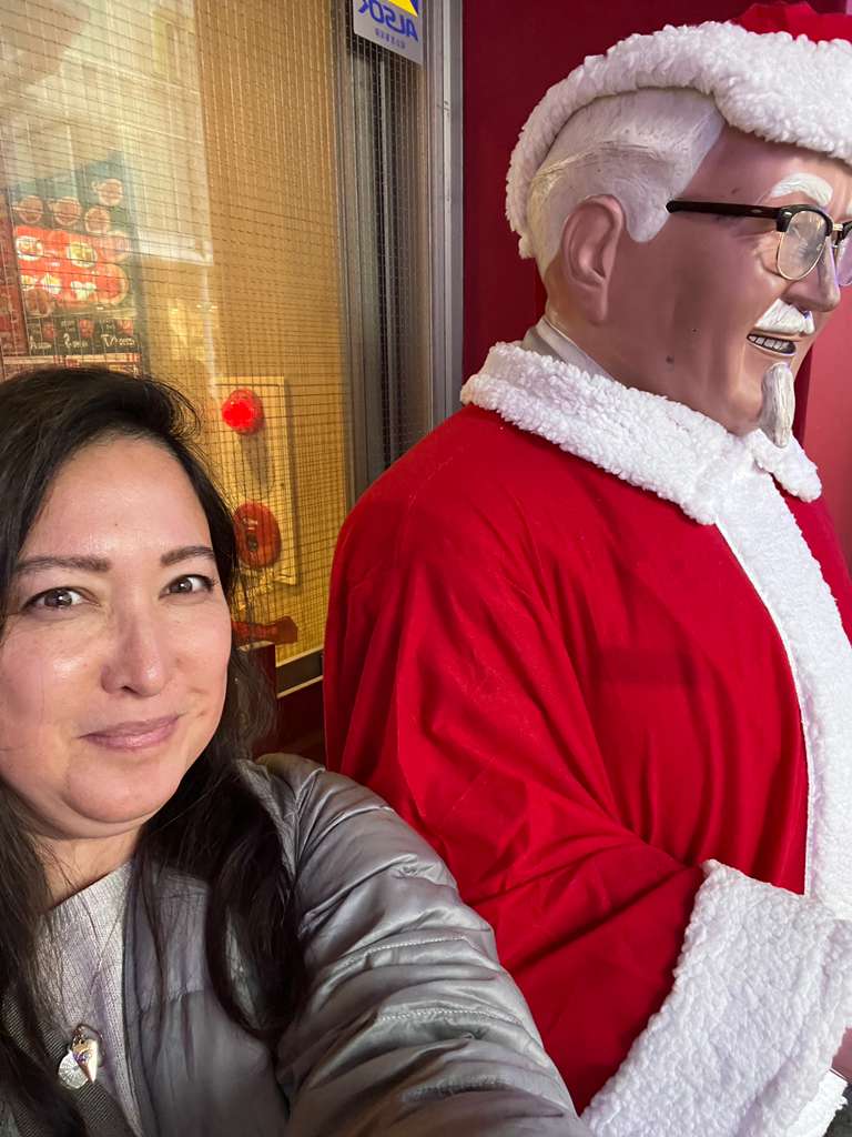 the author Malia Yoshioka with Colonel Sanders statue dressed as Santa claus outside of KFC in Kyoto, Japan in December 2025