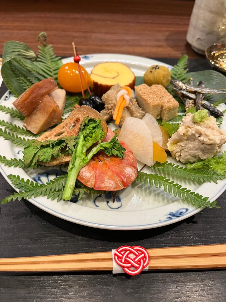 An elegant plate of osechi ryori (new year's foods) at Choshoku Kishin in Gion, Kyoto, served specially for new years in the first days of the year. Each item has a special meaning, including shrimp (longevity), black beans (kuru mame - for studies), karasumi (bottarga), lotus root, loquat, dried sardines, and more, displayed on a bed of green ferns. 