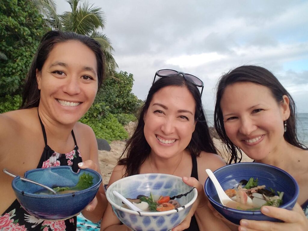 Author Malia Yoshioka having ozoni with two friends at the beach on New year's Day in Hawaii. Palm trees line the beach and the ocean is visible on the horizon, each girl carries a bowl of ozoni soup to hold up to the camera.