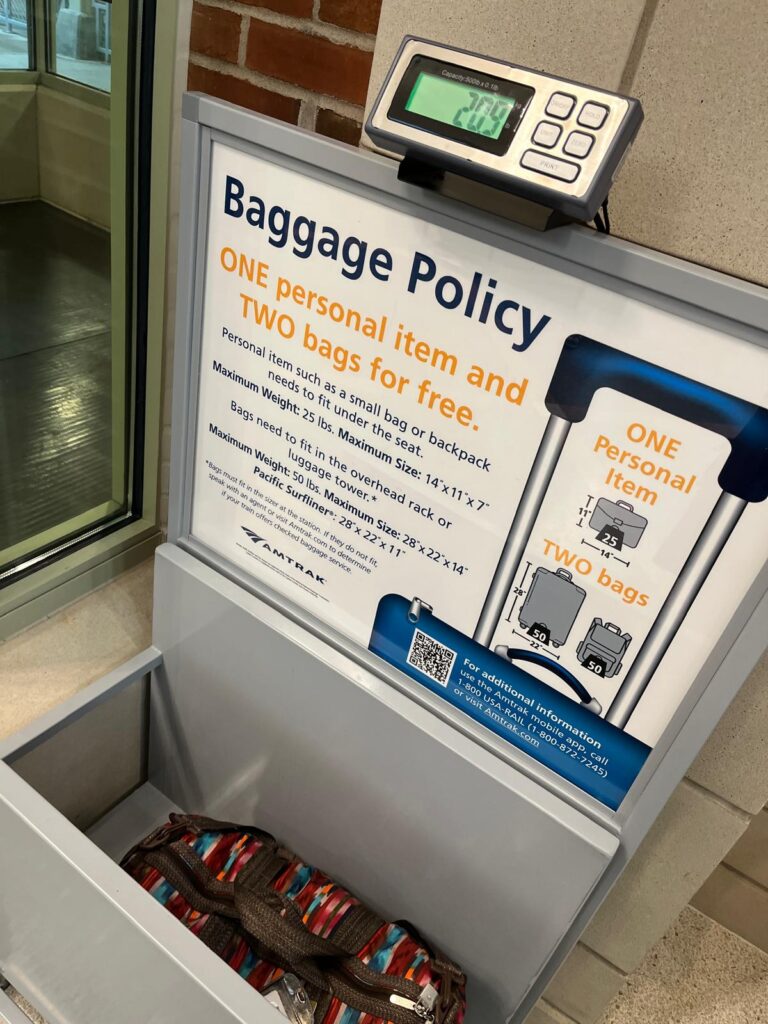 Amtrak baggage policy sign beside a luggage sizer with a suitcase inside. The sign reads "Baggage Policy. ONE personal item and TWO bags for free." It also lists "Personal item such as a small bag or backpack needs to fit under the seat. Maximum Weight 25 lbs. Maximum Size 14 x 11 x 7. Bags need to fit in the overhead rack or luggage tower. Maximum Weight 50 lbs. Maximum Size 28 x 22 x 14." An illustration shows one 25-pound personal item and two 50-pound bags.