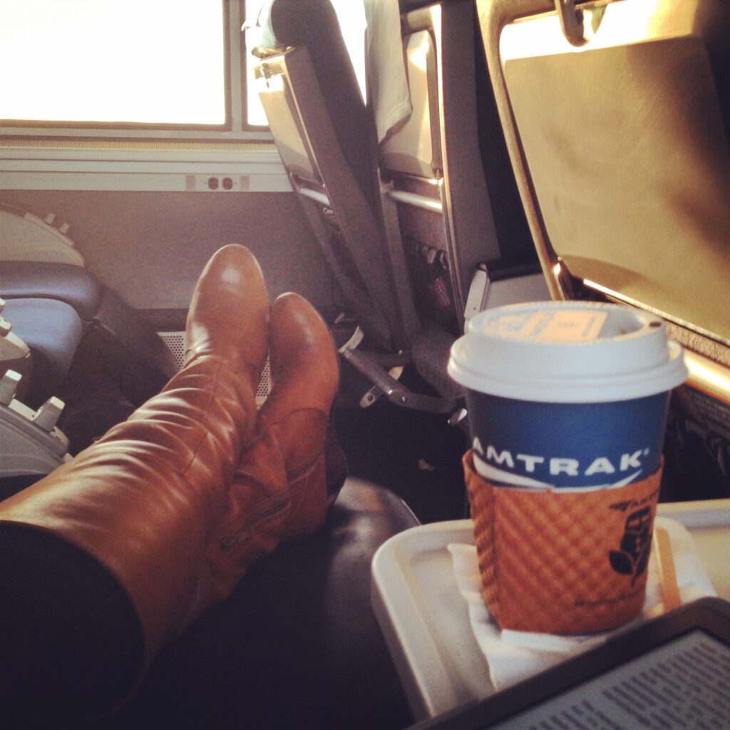 Passenger relaxing in a spacious Amtrak coach seat with boots propped up, a Kindle and a cup of coffee on the tray table beside the window. The photo illustrates the extra legroom available during long-distance train travel in Coach in Amtrak.