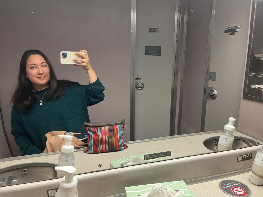 Author Malia Yoshioka taking a mirror selfie inside a spacious Amtrak train restroom with a toiletry bag on the counter beside soap and tissues and two sinks, it is the "Ladies Lounge" cabin that I found on one long distance train route (not available in all trains). The image accompanies tips for staying fresh during overnight train travel.