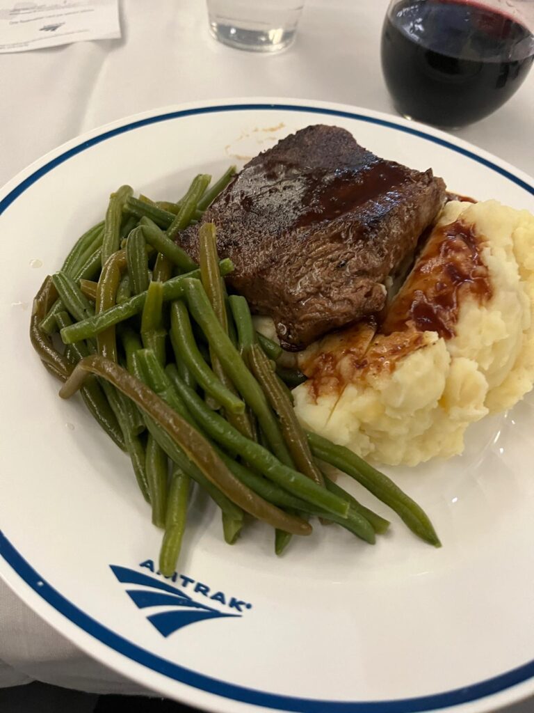 Amtrak dining car dinner with steak, mashed potatoes, green beans, and a glass of red wine on a white plate with the Amtrak logo. The meal illustrates the type of dining available on some long-distance routes.