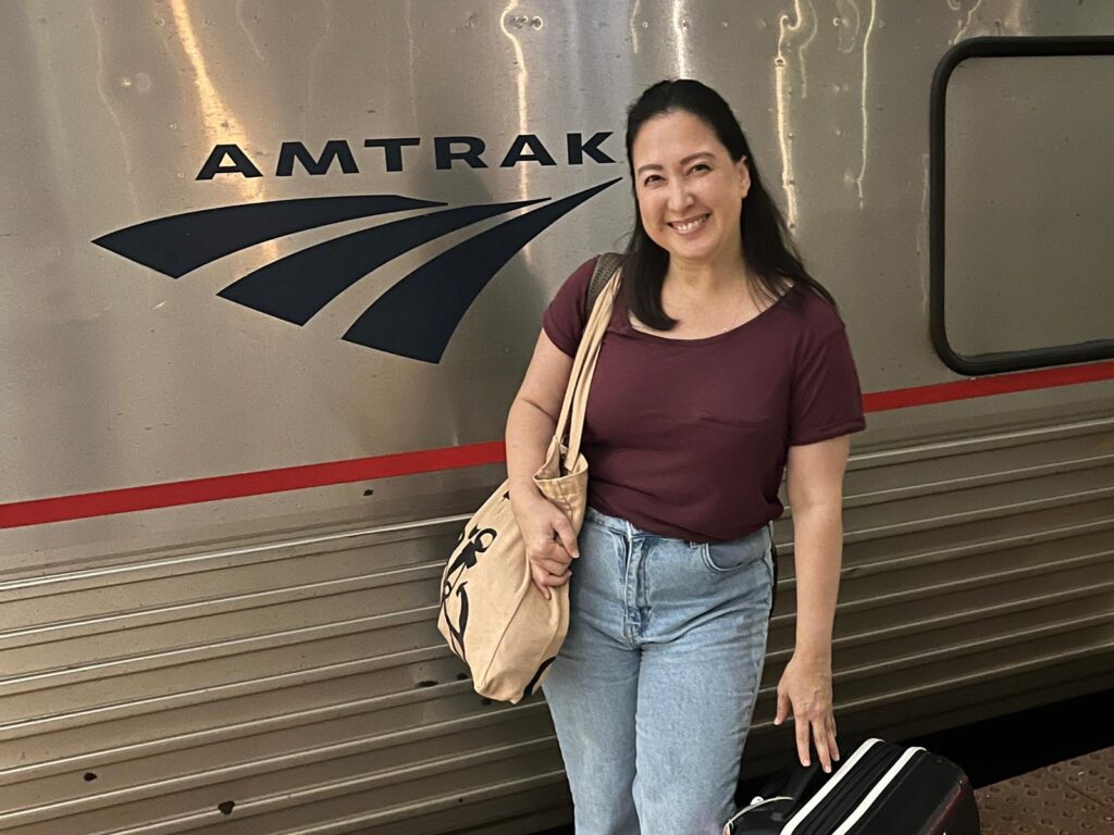 Author Malia Yoshioka smiling beside an Amtrak train with the Amtrak logo visible behind her while holding a tote bag and rolling suitcase. This photo introduces the Amtrak FAQs guide for first-time train travelers.
