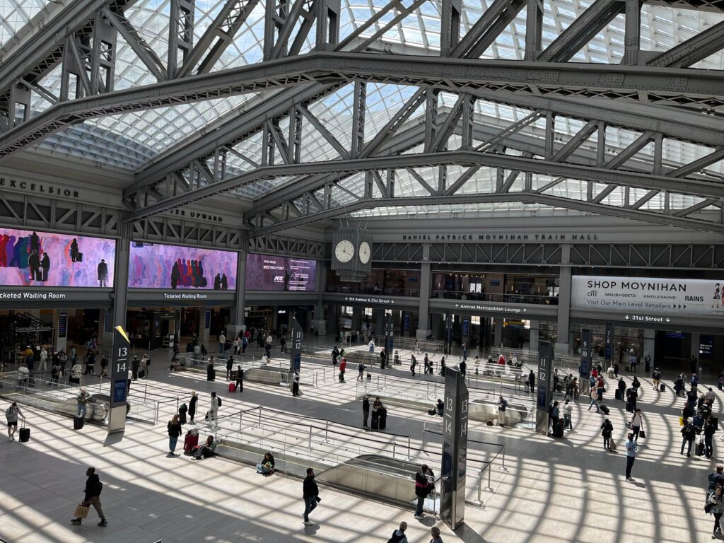 Wide view inside Moynihan Train Hall with travelers walking beneath a glass ceiling and digital departure boards. The photo shows the type of large station first-time Amtrak passengers may encounter.