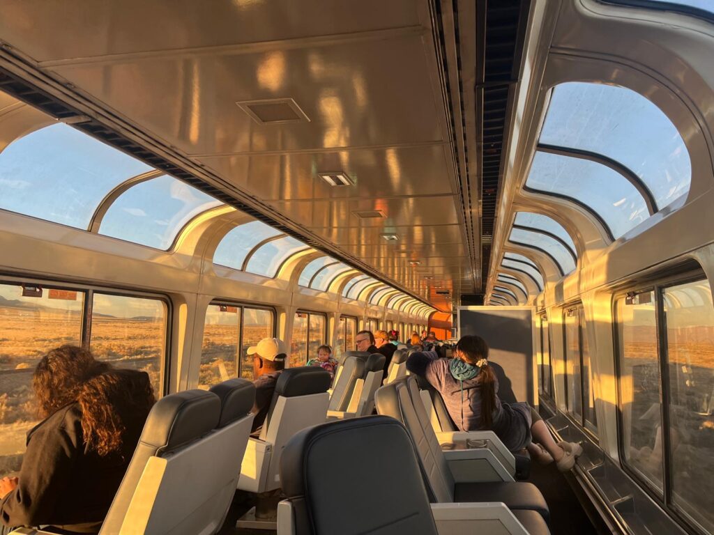 Passengers seated inside an Amtrak Sightseer Lounge surrounded by tall panoramic windows overlooking a sunlit landscape. The photo highlights one of the most scenic spaces and Author Malia Yoshioka's favorite spot described in the Amtrak FAQs guide.