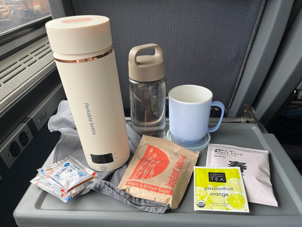 Travel tray with a portable kettle, reusable water bottle, mug, instant oatmeal, tea, coffee packets, and condiments on an Amtrak seat tray. The image shows supplies for preparing simple meals and drinks during a long train journey.